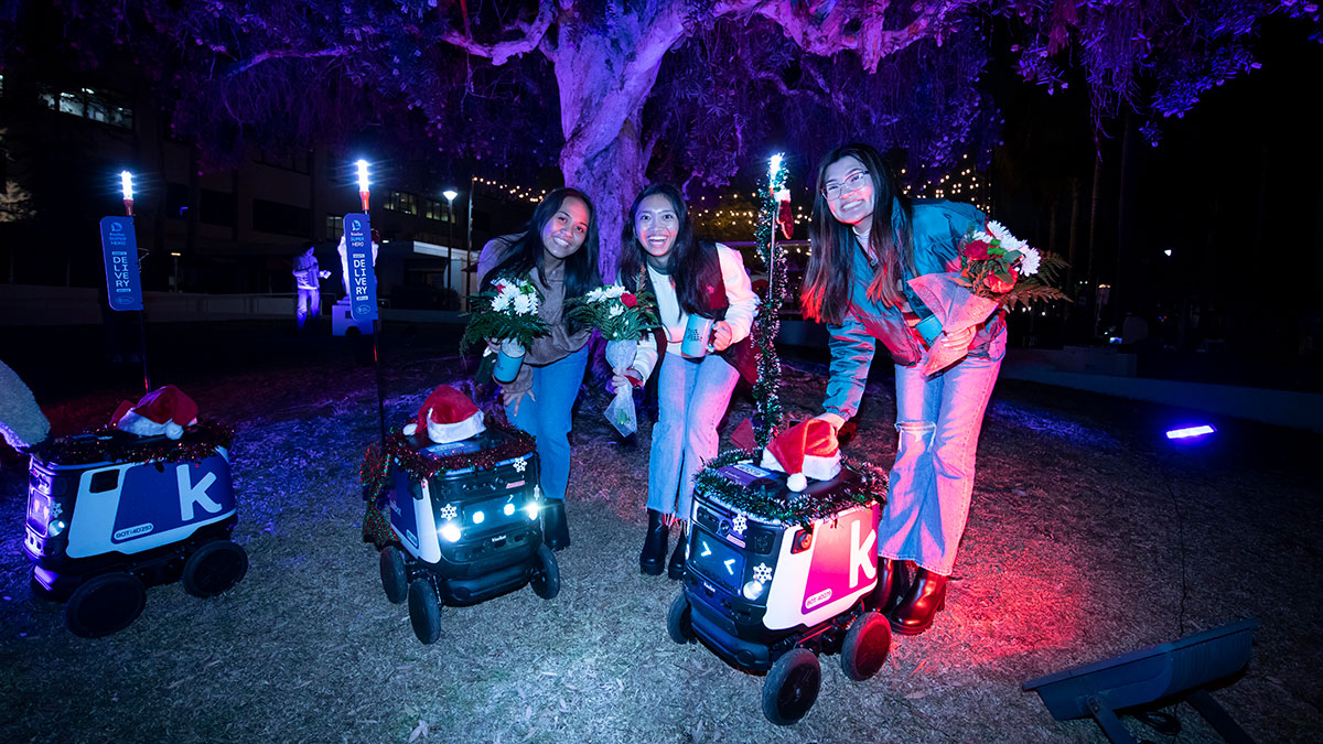 Several students standing with on-campus delivery robots which are decorated with Santa hats