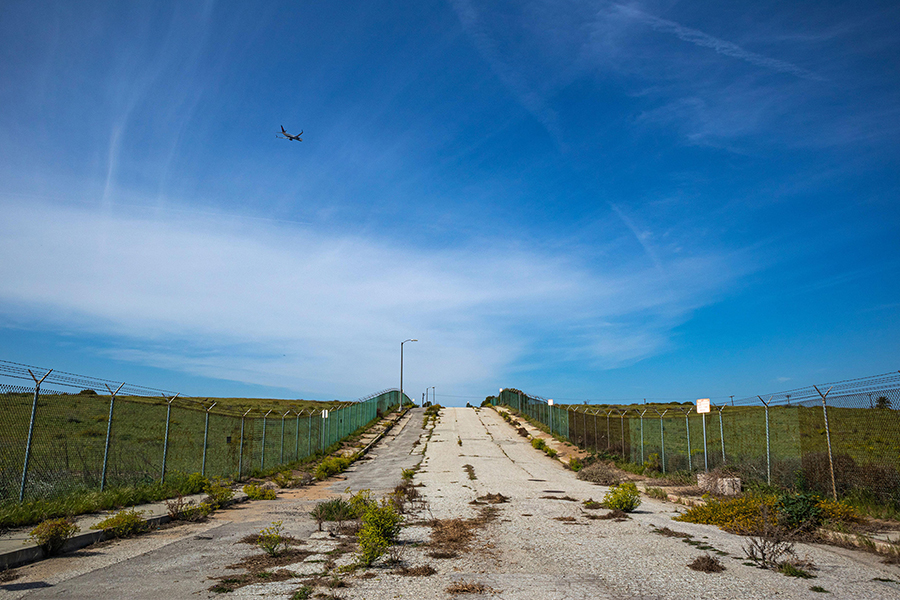 An old paved road with weeds growing in it, & an airplane taking off in the sky