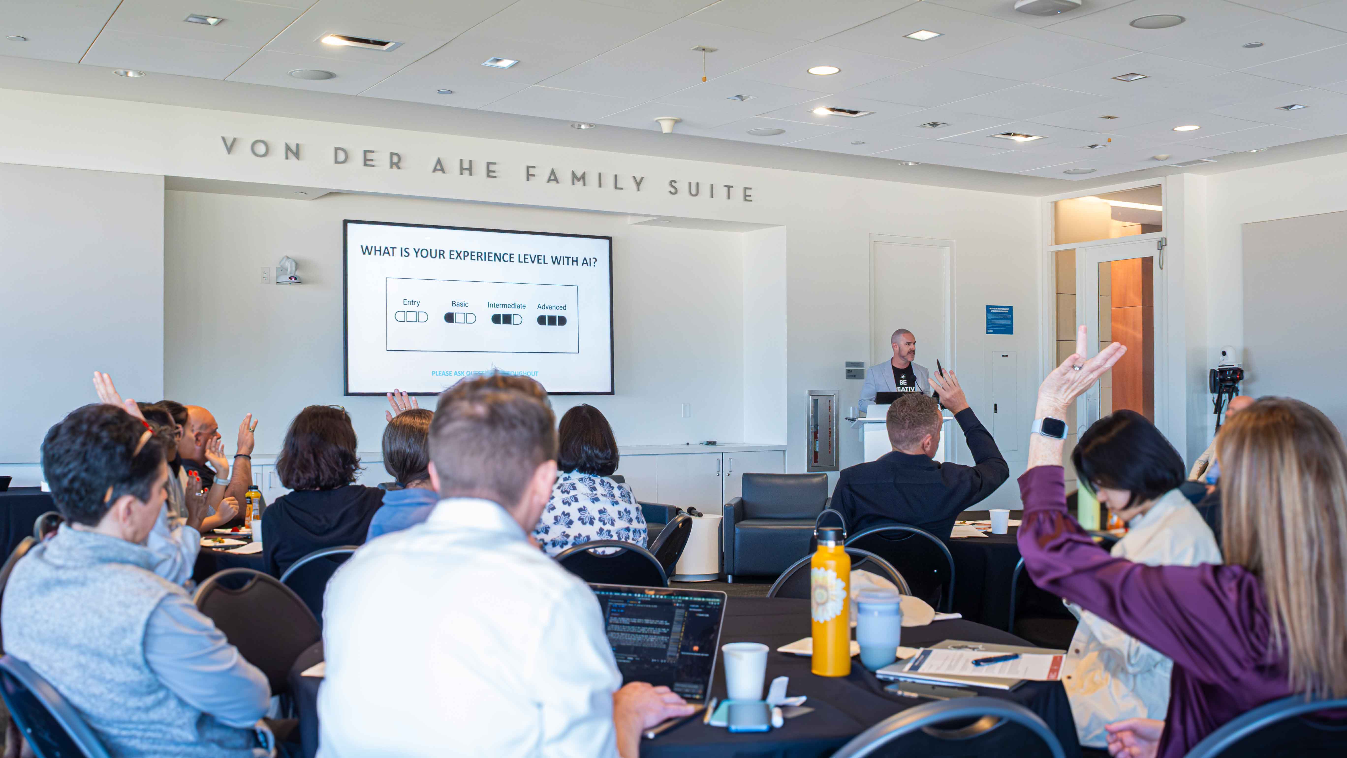 Audience members raise their hands during a presentation in the Von der Ahe Family Suite; a screen asks about their experience level with AI.