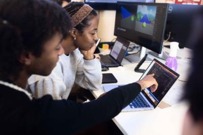 Three people collaborate around laptops and monitors, with one person pointing to code on a laptop screen.