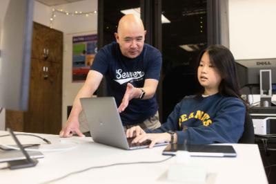 Two people collaborate on laptops at a table, with one person gesturing toward the other’s computer.