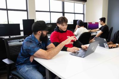 People collaborate around laptops and desktop computers in a bright computer lab.