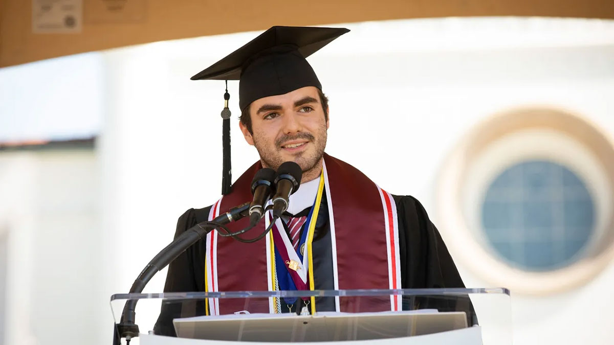 Alfredo Yei Hernandez speaking on stage while wearing a graduation gown