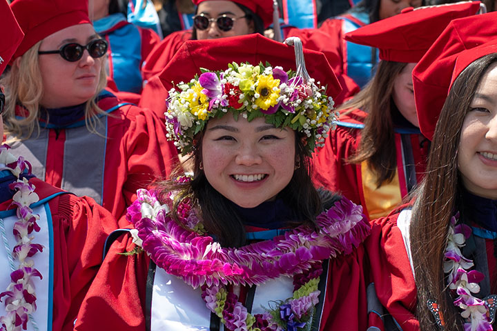 A graduate in cap and gown while sporting a flower crown and two leis during commencement